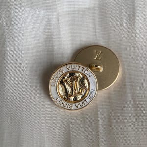 Pode incluir: Dois botões Louis Vuitton dourados. Um botão está virado para cima, exibindo o nome e o logotipo da marca em dourado sobre um fundo branco. O outro botão está virado para baixo, mostrando a parte de trás do botão com o logotipo da marca.