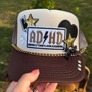 May include: A brown and cream trucker hat featuring a central patch with the text AD/HD HIGHWAYTOHEYLOOKASQUIRREL, accented by gold sequin stars, a white flower, a black heart, a gold chain trim, a small black bow, and three clear rhinestones.