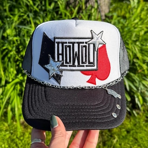 May include: Black and white trucker hat with the word "HOWDY" in a bold font. The hat features a star and spade design, and a silver chain detail. The hat is held by a hand with green nail polish.