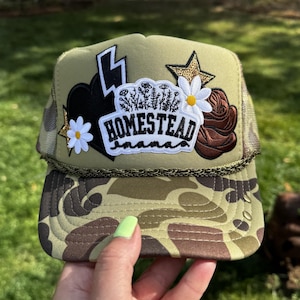 May include: An olive green trucker hat featuring a camouflage brim and a central patch with the text HOMESTEAD mama. The design includes embroidered white daisies, a black heart, a lightning bolt, a gold sequin star, and a brown rose patch.