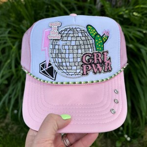 May include: Pink and white trucker hat with a disco ball, cactus, and diamond patch design. The hat has the text "GRL PWR" and "smile to the world". Rhinestones and green beading adorn the brim.