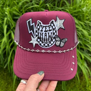 May include: Burgundy trucker hat with a mesh back and a curved brim. The front features a lightning bolt, star, horseshoe, and butterfly embellishments. The text "YEE HAW" is displayed in a checkered pattern.