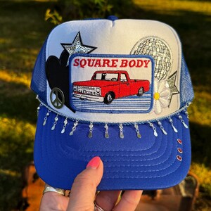 May include: A blue and white trucker hat featuring a central rectangular patch with a red pickup truck and the text SQUARE BODY. The hat is decorated with a silver peace sign, a sequined star, a disco ball patch, a white flower, and dangling silver charms.