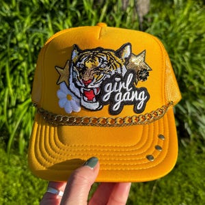 May include: Mustard yellow trucker hat with a tiger patch, gold chain, and the words "girl gang". The hat has a mesh back and a curved brim. The tiger patch includes a white daisy and gold stars.