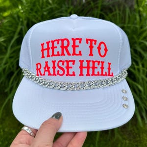 May include: White trucker hat with the text "HERE TO RAISE HELL" in red. The hat features a silver chain detail and three silver studs on the brim. The hat is held up by a hand.