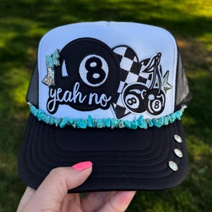 May include: A white and black trucker hat featuring a black patch with an 8-ball and the text yeah no, a checkered heart, and cherry graphics. The brim is black with three silver studs and a turquoise stone beaded trim across the front base.