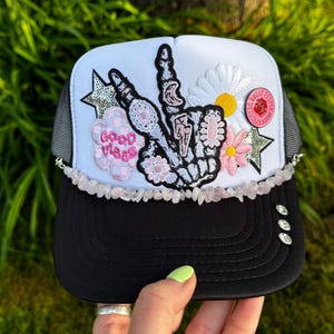 Puede incluir: Gorra de camionero en blanco y negro con la parte trasera de malla. La parte delantera presenta un signo de la paz con un diseño de esqueleto, flores, estrellas y las palabras "Good Vibes". La visera es negra y el sombrero está decorado con pequeñas piedras.