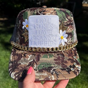 May include: A camouflage trucker hat featuring a white rectangular patch with the text "HOT GIRLS HAVE GRANNY HOBBIES" in white lettering. The hat is decorated with two white daisy appliques, two gold glitter stars, a gold chain, and rhinestones.