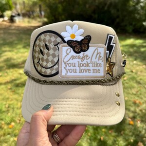 May include: A beige trucker hat featuring a checkered smiley face patch, a white daisy, a butterfly, a black lightning bolt, and a gold star. A rectangular patch reads, "Excuse Me, you look like you love me." A thin gold chain accents the brim.