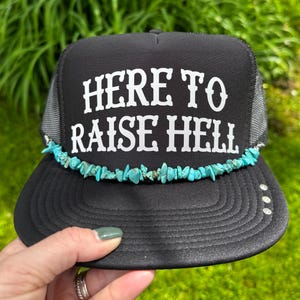 May include: Black trucker hat with the text "HERE TO RAISE HELL" in white. The hat features a turquoise stone embellishment along the brim. The hat has a black brim and a gray mesh back.