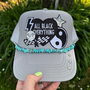 May include: Gray trucker hat with a mesh back and a curved brim. The front features patches with the text "ALL BLACK EVERYTHING", a lightning bolt, a yin-yang symbol, and a star. A turquoise stone bead trim accents the brim.