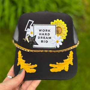 May include: Black trucker hat with a mesh back, featuring a white square with the text "WORK HARD DREAM BIG". Embellished with a sunflower, daisies, a gold chain, and yellow embroidered accents. A lightning bolt and star add to the design.