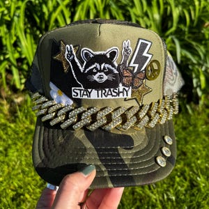 Puede incluir: Gorra de camionero de camuflaje con una cadena de pedrería y parches. Los parches incluyen un mapache, un rayo, un símbolo de la paz y las palabras "Stay Trashy". La gorra tiene una parte trasera de malla y una visera curva.
