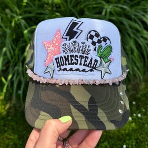 May include: A camouflage trucker hat with a white front panel featuring a pink star, lightning bolt, and a black and white checkered heart. The hat has the text "HOMESTEAD mama" and is trimmed with pink beads.