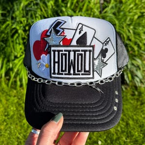 May include: Black and white trucker hat with a chain and decorative accents. The hat features a white front panel with playing card and star designs, the word "HOWDY" in a rectangular box, and a black brim.