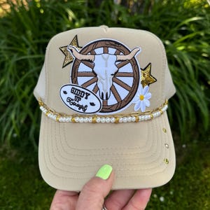 May include: Tan trucker hat with a bull skull and wagon wheel patch. The patch is surrounded by gold stars and a daisy. The brim is tan with gold eyelets. The hat has a pearl and gold chain around the brim and the text "Giddy Up Cowgirl".