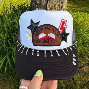 May include: A black and white trucker hat with a mesh back. The front features a patch with the text "SHOW ME YOUR BOBBERS", a lightning bolt, stars, and a daisy. The brim is decorated with dangling silver charms.