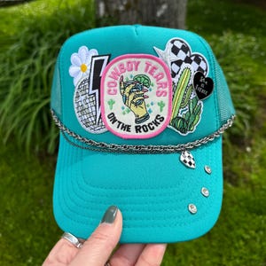 May include: A turquoise trucker hat with a mesh back and a curved brim. The hat is decorated with patches, including a disco ball, a daisy, and a patch that says "Cowboy Tears on the Rocks". A silver chain and rhinestones add detail.