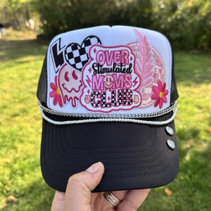 May include: A white and black trucker hat featuring a central pink patch with the text OVER STIMULATED MOMS CLUB, a checkered heart, a melting smiley face, pink flowers, and a feather. The brim is black with two silver studs and a chain trim.