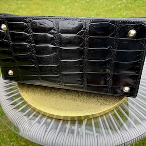 May include: A rectangular black handbag with a glossy crocodile-embossed texture. The base features four polished silver-tone metal feet at each corner, providing stability and protection for the bottom surface of the bag.