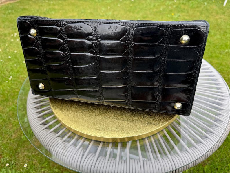 May include: A rectangular black handbag with a glossy crocodile-embossed texture. The base features four polished silver-tone metal feet at each corner, providing stability and protection for the bottom surface of the bag.
