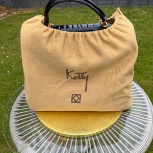 May include: A tan fabric dust bag with a drawstring closure, featuring the word Ketty and a square logo printed in dark brown. The top of a black crocodile-textured handbag with a glossy finish and gold hardware is visible above the bag.