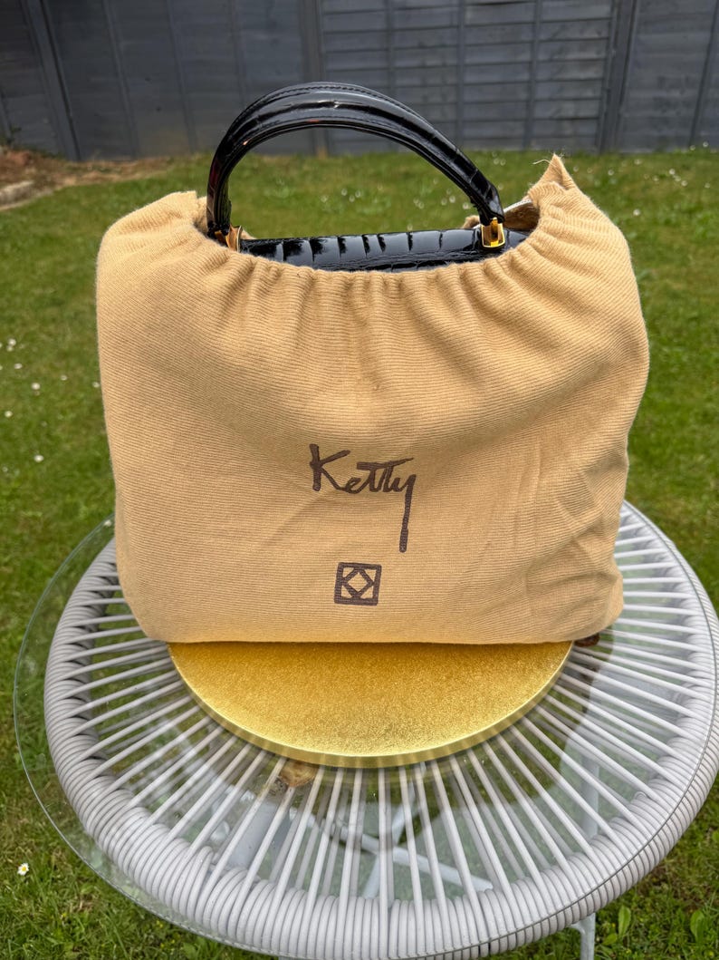 May include: A tan fabric dust bag with a drawstring closure, featuring the word Ketty and a square logo printed in dark brown. The top of a black crocodile-textured handbag with a glossy finish and gold hardware is visible above the bag.