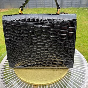 May include: A structured black handbag with a glossy, embossed crocodile skin texture. The bag features a single arched top handle attached with gold-toned metal hardware, a rectangular silhouette, and a flap closure that covers the main compartment.