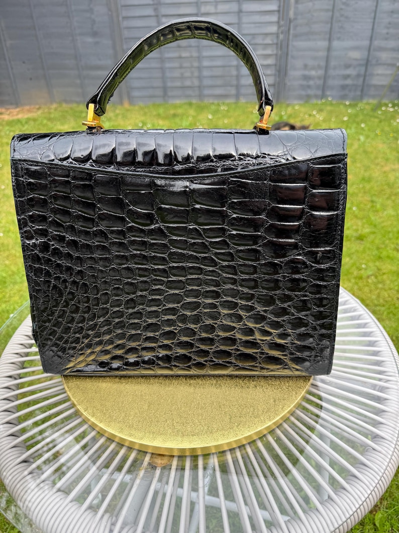 May include: A structured black handbag with a glossy, embossed crocodile skin texture. The bag features a single arched top handle attached with gold-toned metal hardware, a rectangular silhouette, and a flap closure that covers the main compartment.