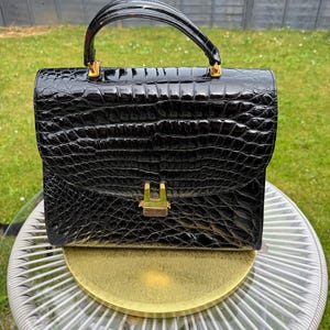 May include: A black handbag with a glossy, textured crocodile-embossed finish. The structured bag features a single arched top handle, a rectangular flap closure, and a gold-toned metal clasp centered on the front. The hardware is polished gold.