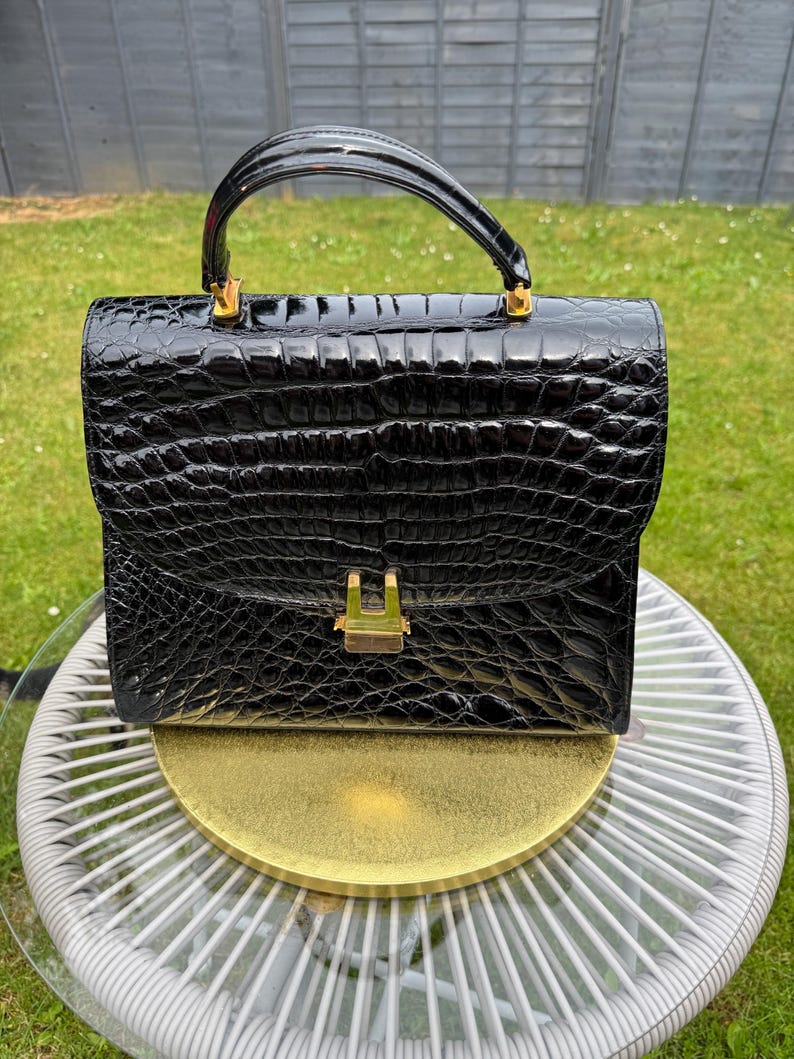May include: A black handbag with a glossy, textured crocodile-embossed finish. The structured bag features a single arched top handle, a rectangular flap closure, and a gold-toned metal clasp centered on the front. The hardware is polished gold.
