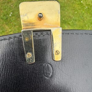 May include: A black leather bag featuring a gold-toned metal clasp mechanism. The rectangular metal hardware is secured with two visible screws and includes a central post. The leather surface shows a subtle embossed circular logo detail.