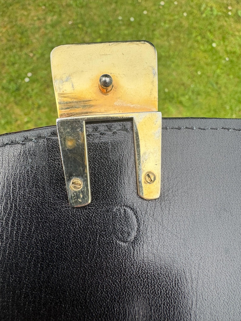 May include: A black leather bag featuring a gold-toned metal clasp mechanism. The rectangular metal hardware is secured with two visible screws and includes a central post. The leather surface shows a subtle embossed circular logo detail.