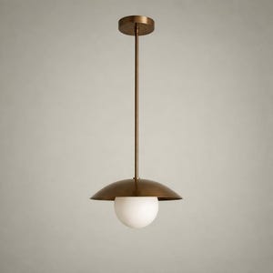 May include: A bronze-colored pendant light fixture with a round canopy and a long, slender rod. The fixture features a downward-facing, saucer-shaped shade and a spherical, white glass globe. The light is suspended against a neutral background.