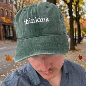 Può includere: Un berretto da baseball verde con la scritta "thinking" ricamata in bianco. Il berretto è indossato da una persona che indossa una camicia blu abbottonata. Lo sfondo mostra una strada cittadina con foglie autunnali che cadono.