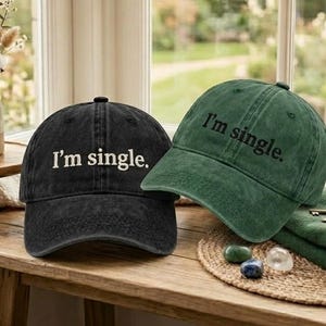 May include: Two baseball caps, one black and one green, each with the text "I'm single" embroidered in white. The caps are displayed on a wooden surface near a window, with a vase of flowers in the background.