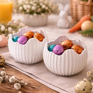 Può includere: Due ciotole in ceramica bianche e scanalate riempite con uova di cioccolato avvolte in carta stagnola nei colori verde acqua, viola e rame. Le ciotole sono su una tavola di legno con un panno di lino neutro, con decorazioni pasquali sullo sfondo.
