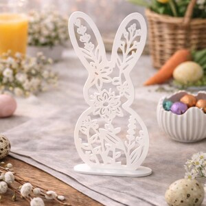 Könnte beinhalten: Eine weiße, lasergeschnittene Osterhasen-Dekoration mit floralen und Blattdetails. Der Hase steht auf einem kleinen, runden Sockel. Der Hintergrund zeigt Ostereier, Karotten und einen Korb mit gelben Blumen, was ein Frühlingsmotiv suggeriert.