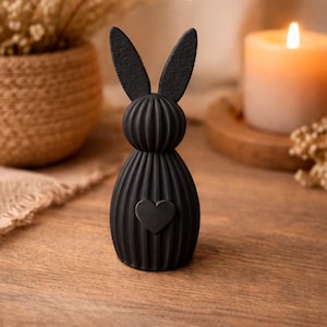Könnte beinhalten: Schwarze Hasenfigur mit Herz-Akzent. Der Hase hat eine gerippte Textur und lange Ohren. Die Figur steht auf einer Holzoberfläche, im Hintergrund eine Kerze und Trockenblumen.
