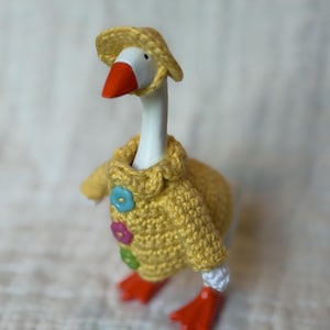 May include: A white goose figurine with an orange beak and feet, wearing a yellow crocheted jumper featuring three flower-shaped buttons in light blue, pink, and green, and a matching yellow crocheted hat perched on its head.