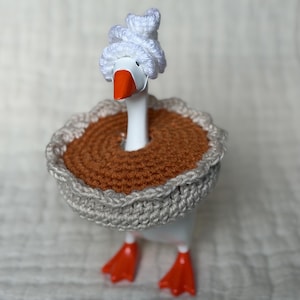 May include: A white plastic goose figurine wearing a crocheted white towel wrap on its head and a crocheted brown and beige pie-shaped collar around its neck. The goose has an orange beak and orange webbed feet.