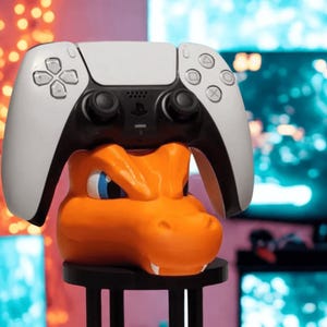 Può includere: Un supporto per controller di videogiochi arancione e nero a forma di drago dei cartoni animati. Un controller di videogiochi bianco e nero poggia sulla parte superiore del supporto. Il supporto ha una base nera con quattro gambe. Lo sfondo è sfocato con monitor di computer.