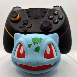 May include: A black video game controller with orange, yellow, and purple accents is positioned above a light blue Bulbasaur figure. The Bulbasaur figure has red eyes and a green design on its head. The items are on a white surface.