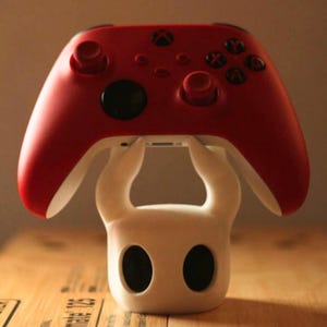 Può includere: Un controller Xbox rosso è appoggiato su un supporto bianco stilizzato. Il controller ha pulsanti neri e un lato inferiore bianco. Il supporto presenta un design con due grandi aperture nere a forma di occhi e una forma curva.