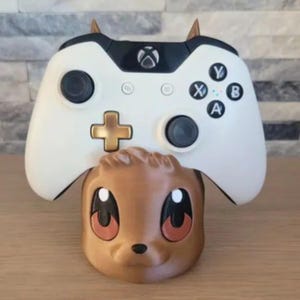 Puede incluir: Un mando de Xbox blanco y negro descansa sobre un soporte de personaje de dibujos animados marrón. El mando tiene botones negros y una cruceta dorada. El soporte tiene ojos grandes y cuernos pequeños.
