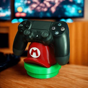 Puede incluir: Un mando negro de PlayStation 4 descansa sobre un accesorio de juego rojo y verde. El accesorio tiene forma de sombrero de Mario con una "M" blanca en la parte delantera, sobre una base verde. El fondo está borroso, mostrando una pantalla de televisión.