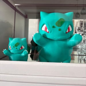 May include: Two teal Bulbasaur figures are displayed, one large and one small. The larger figure has outstretched arms and red and white eyes. Both figures have a green patch on their foreheads and white claws.