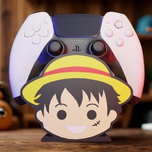 May include: A white and black PlayStation 5 controller with a cartoon character stand featuring a yellow hat and a smiling face. The stand is placed in front of the controller.