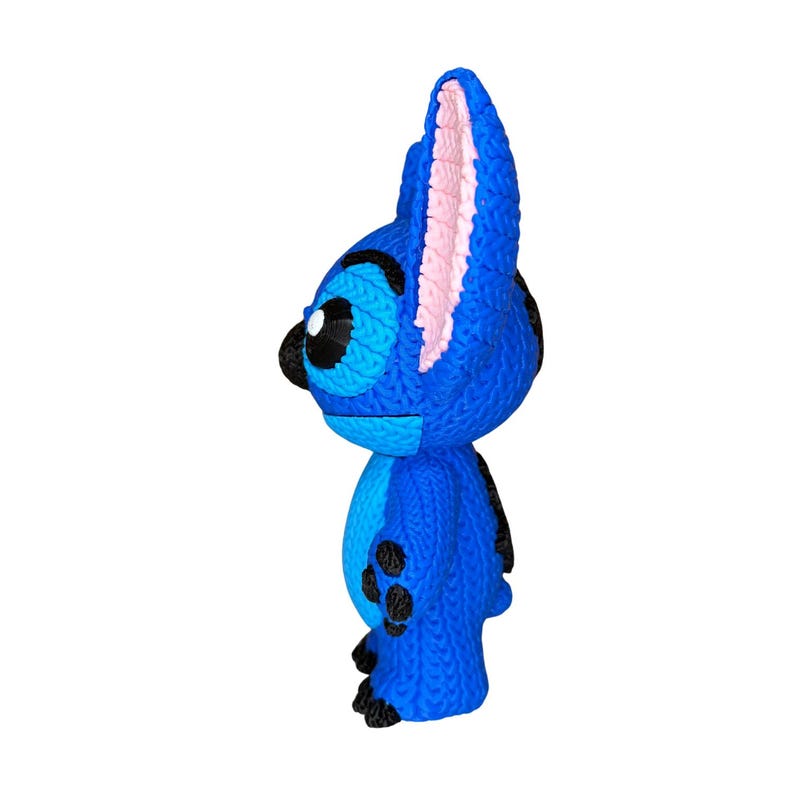 May include: A blue figurine of a cartoon character with large ears and black spots. The figurine has a knitted texture, with pink inner ears and a black nose and eyes. The character is standing upright against a white background.