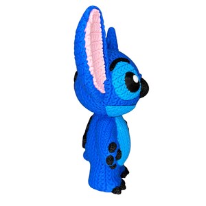 May include: A blue figurine of the character Stitch from the movie Lilo & Stitch. The figurine has large ears with pink inner ears, black eyes, and black spots on its body. The figurine is made of a textured material.